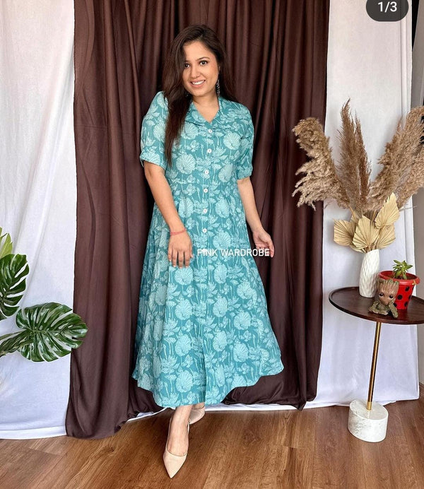 Woman wearing blue floral cotton dress standing by curtain in casual indoor setting
