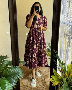 Woman wearing a purple floral maxi dress with sleeves standing in front of a mirror with green plants around